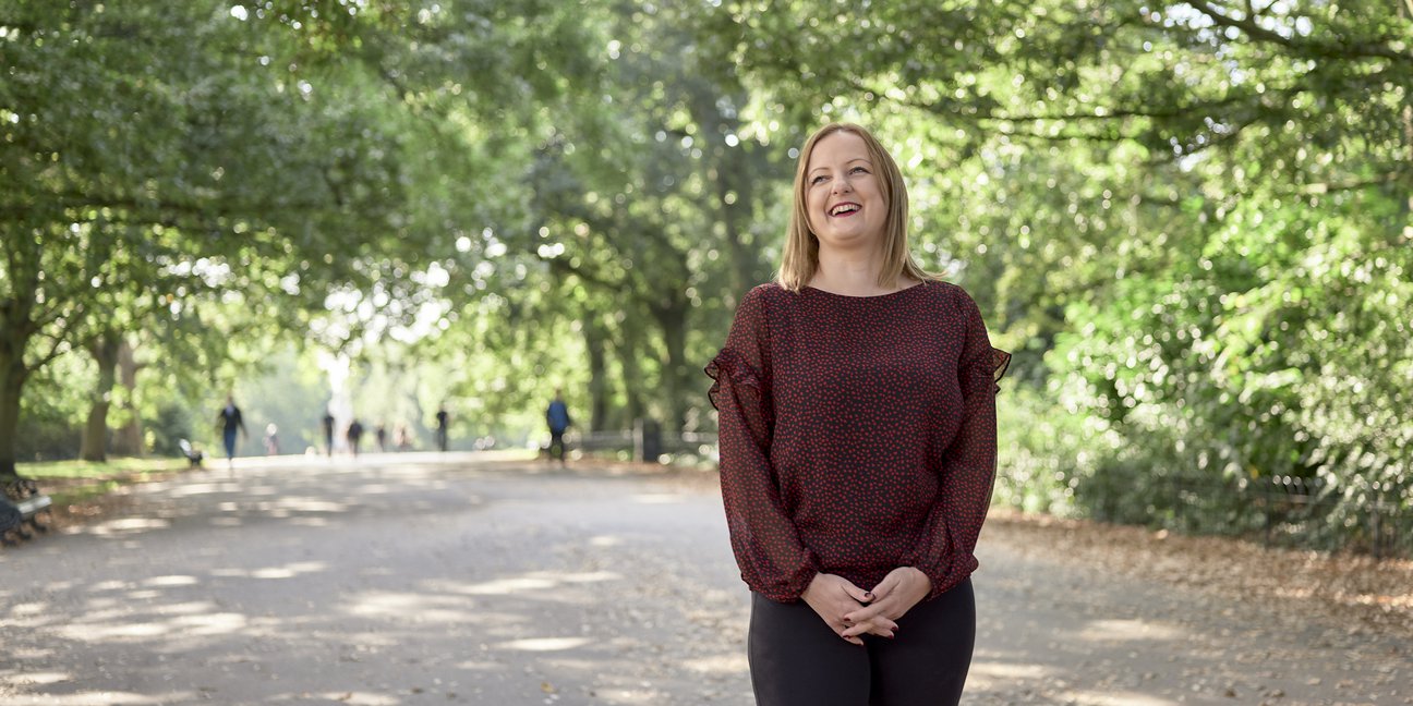 A woman smiles in a park, in front of a backdrop of trees, with dappled light shining through.