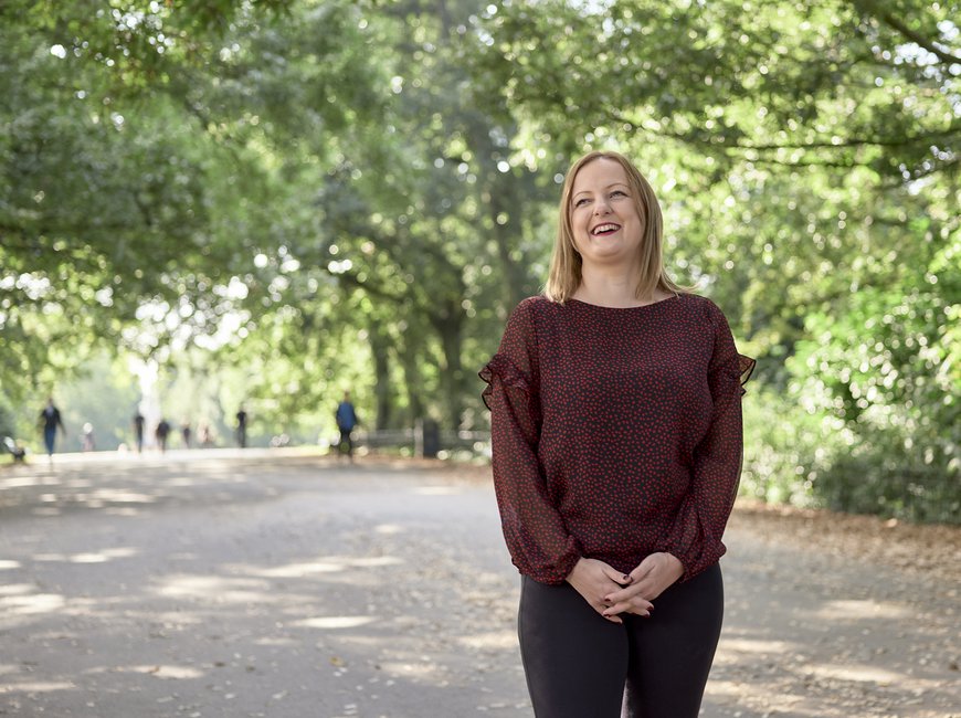 A woman smiles in a park, in front of a backdrop of trees, with dappled light shining through.