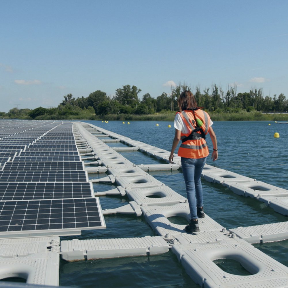 Mreža solarnih panela pluta na mirnom, plavom jezeru pod suncem. Žena u prsluku za spašavanje pažljivo korača stazom na napuhavanje pokraj njih.