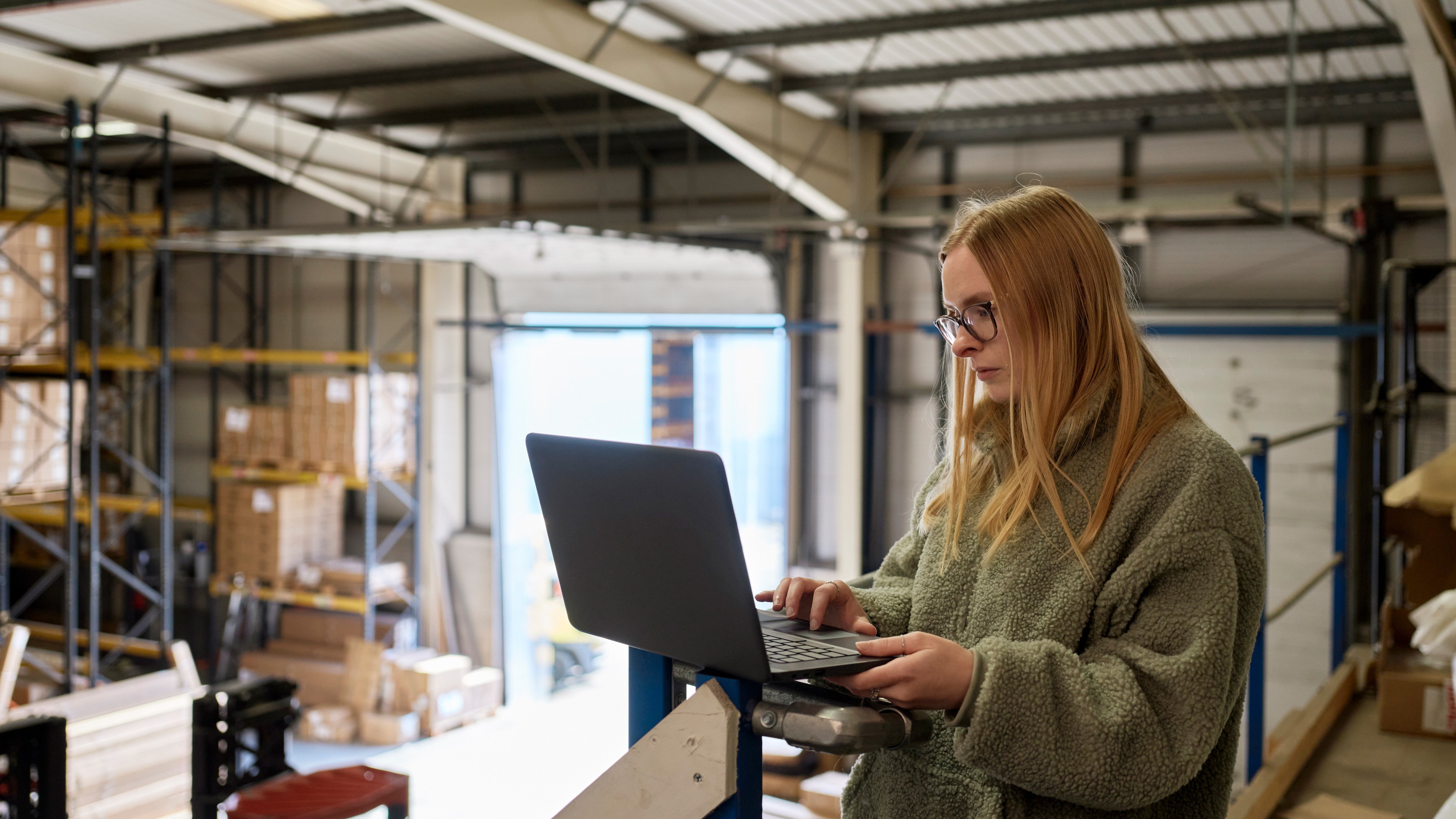 Eine Frau in einer Fleecejacke arbeitet in einer Lagerhalle an ihrem Laptop.