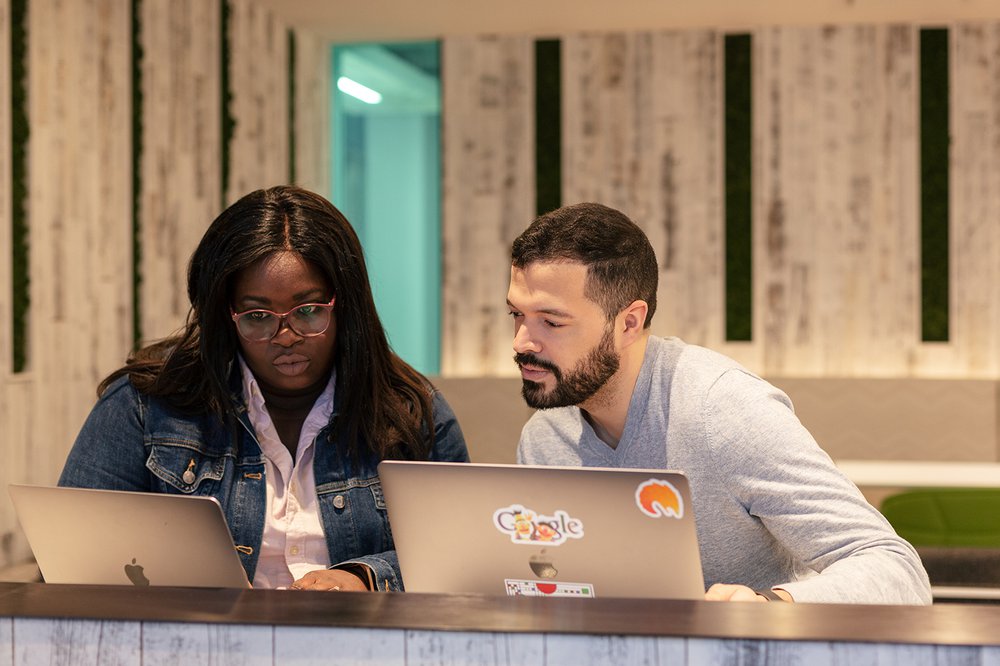 A man and a woman sit at a breakfast bar with their laptops open. Bright stickers are visible on the lid of the man’s laptop.