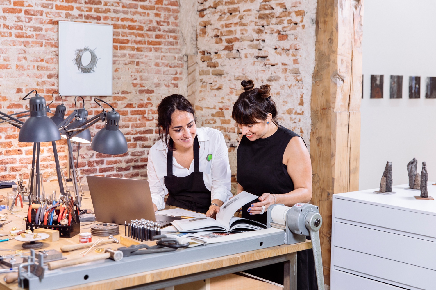 Dvojica žien v dielni na výrobu šperkov si spoločne prezerá príručku a notebook. Sedia pri pracovnom stole obloženom kladivkami, drôtmi, kliešťami a jasnými lampami namierenými k pracovným stolom.