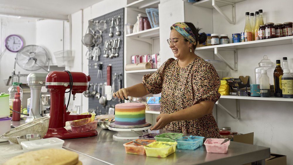 Nainen värikkäässä topissa ja huivi päässään kuorruttaa kakkua leipomossaan. Hänen takanaan seinällä on leipomistarvikkeita, jauhoja ja pakkausmateriaaleja.