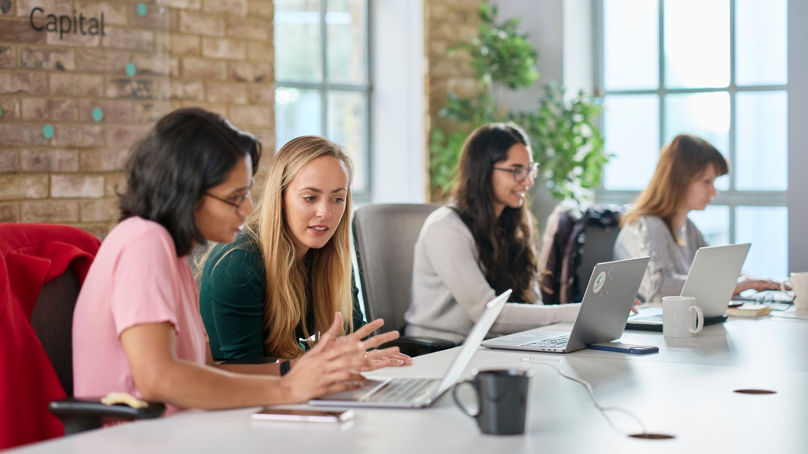 A group of colleagues are sat in a workspace discussing points from their laptop screens.