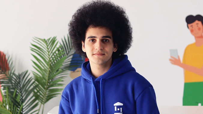 Bir Afro saçlı öğrenci, mavi renkte bir akademi sweatshirt'ü giymiş şekilde gülümseyerek fotoğraf çektiriyor.