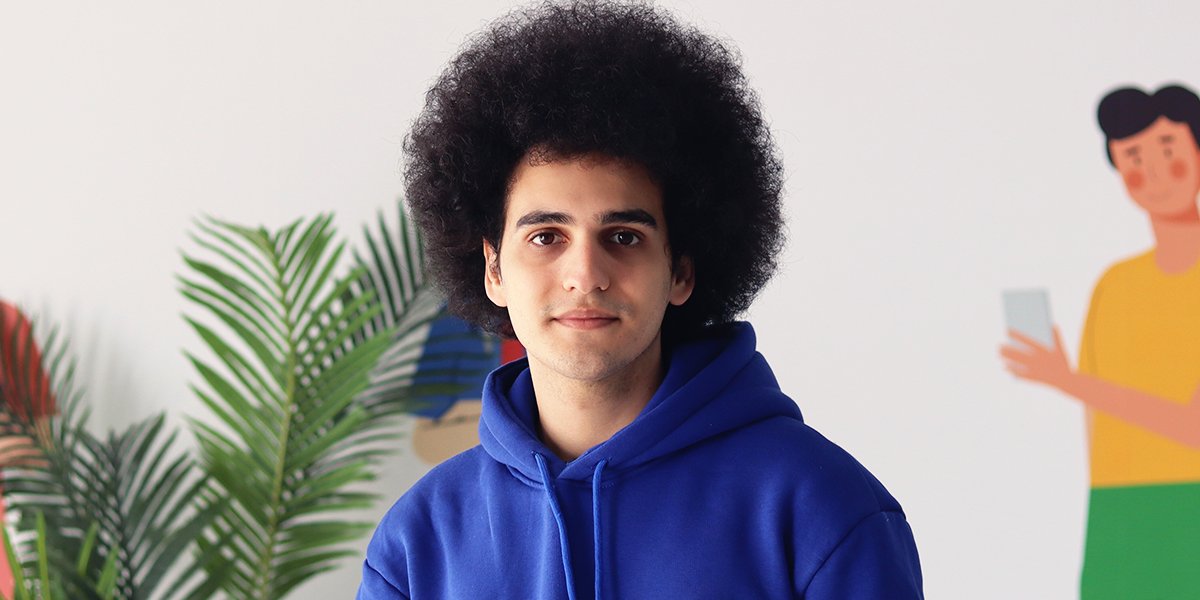 Bir Afro saçlı öğrenci, mavi renkte bir akademi sweatshirt'ü giymiş şekilde gülümseyerek fotoğraf çektiriyor.