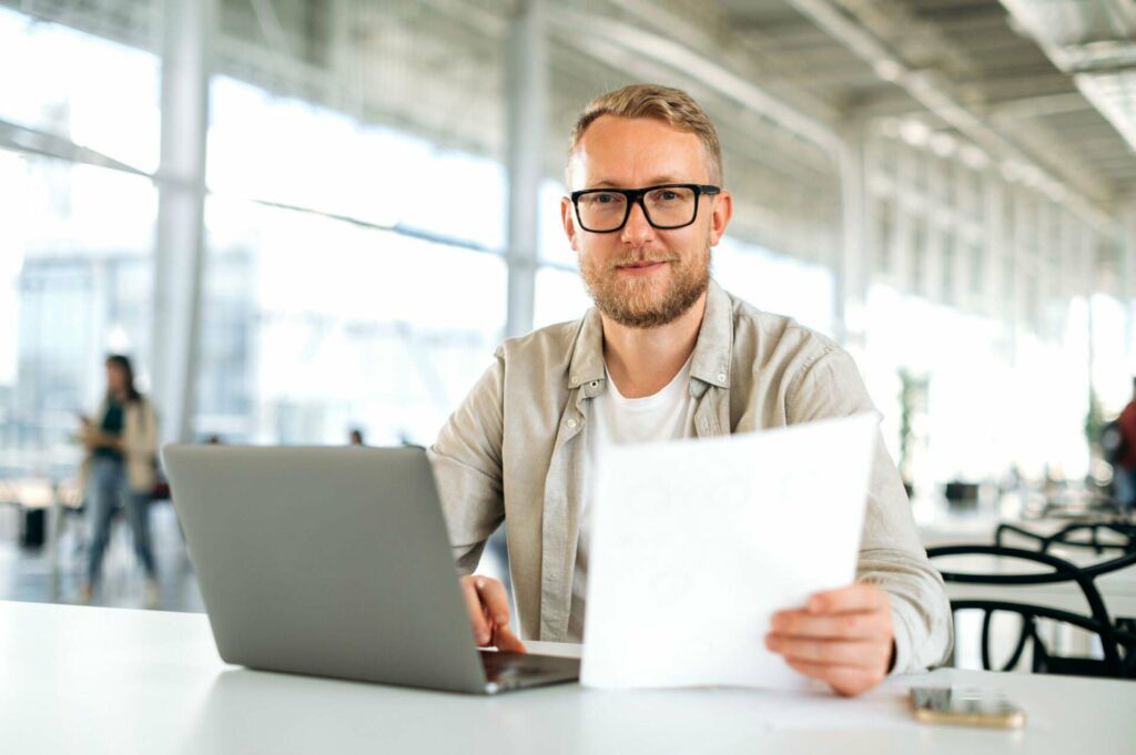 Friendly motivated successful caucasian man, with glasses, IT specialist, company seo, manager, freelancer, sits at a desk with a laptop in coworking, works with documents, looks at camera, smiles Montana