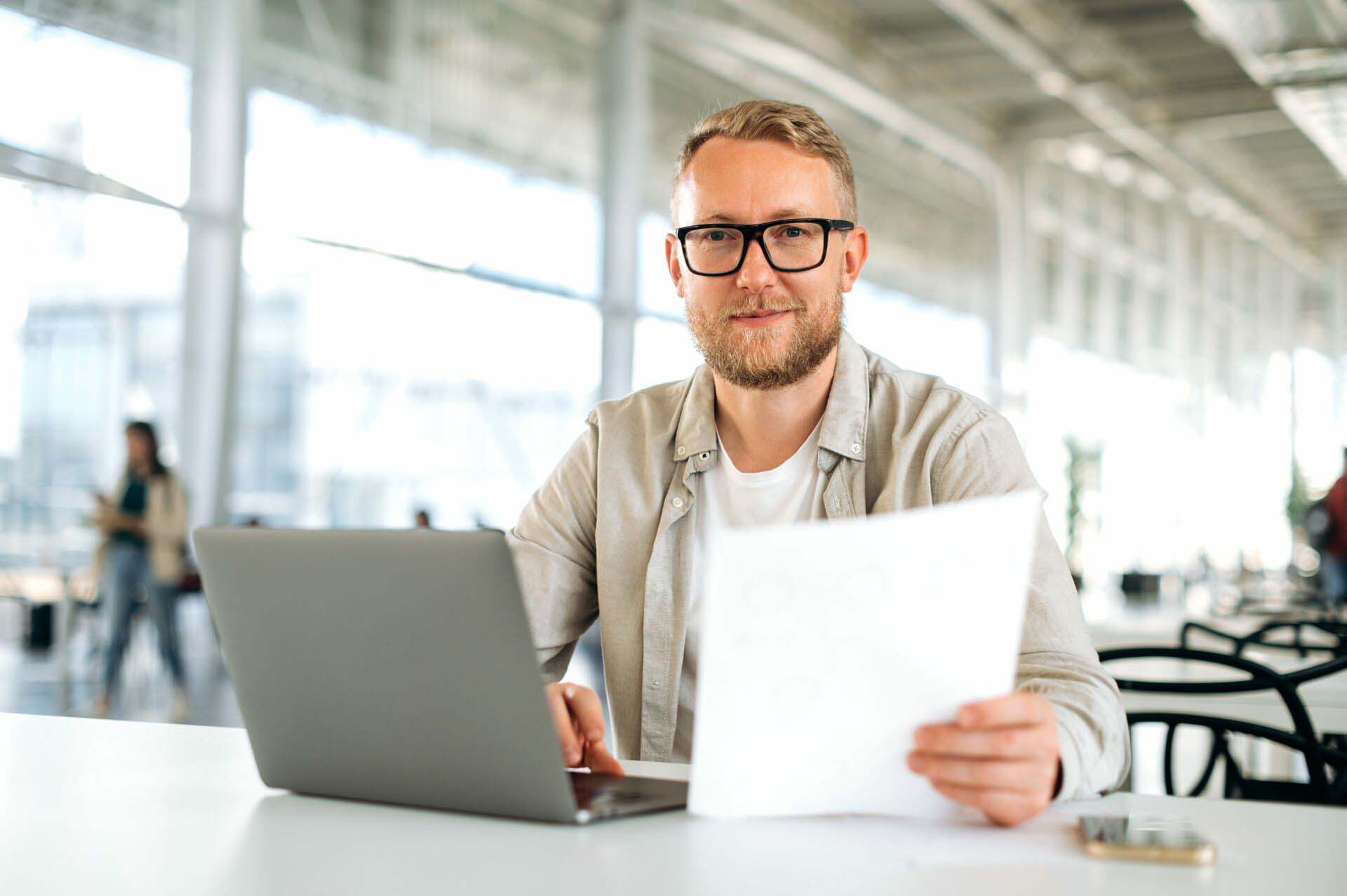 Friendly motivated successful caucasian man, with glasses, IT specialist, company seo, manager, freelancer, sits at a desk with a laptop in coworking, works with documents, looks at camera, smiles Michigan