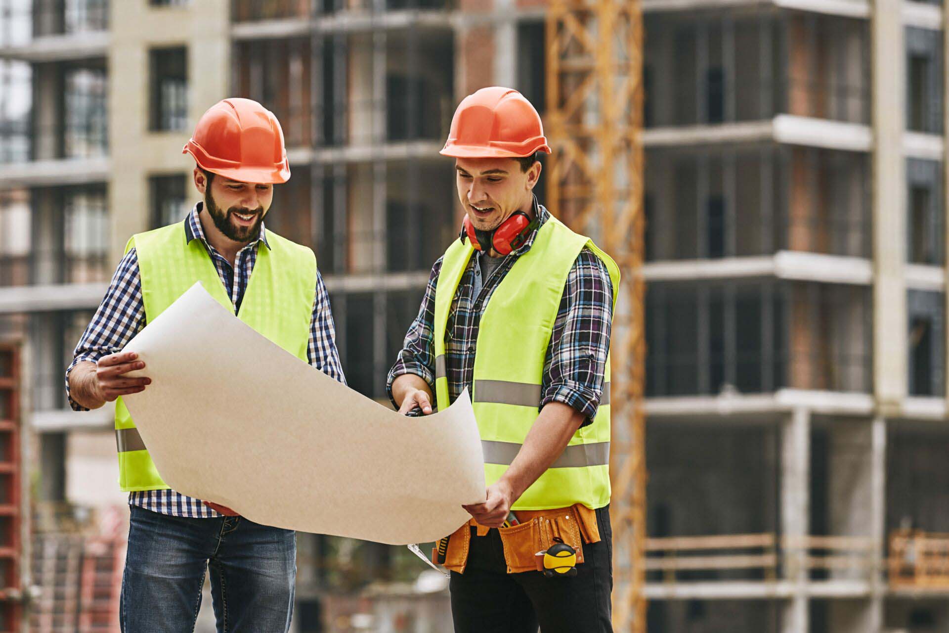 Look here! Two young builders in working uniform and helmets are holding a construction drawing in their hands and discussing it with each other while standing at construction site. Insurance for Construction Project Managers