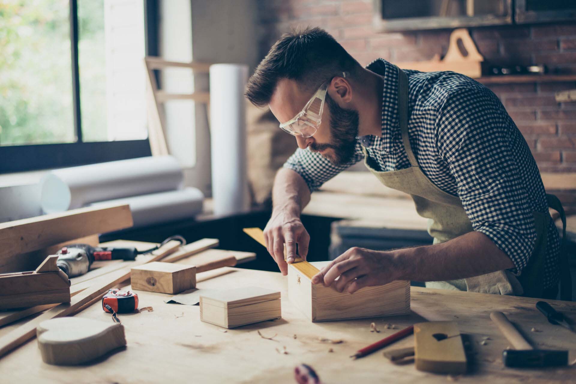 Concentrated persistent talented professional carpenter bearded wearing casual checkered shirt apron safety glasses is measuring the length on wooden box’s side using a ruler Insurance for Crafters and Artisans