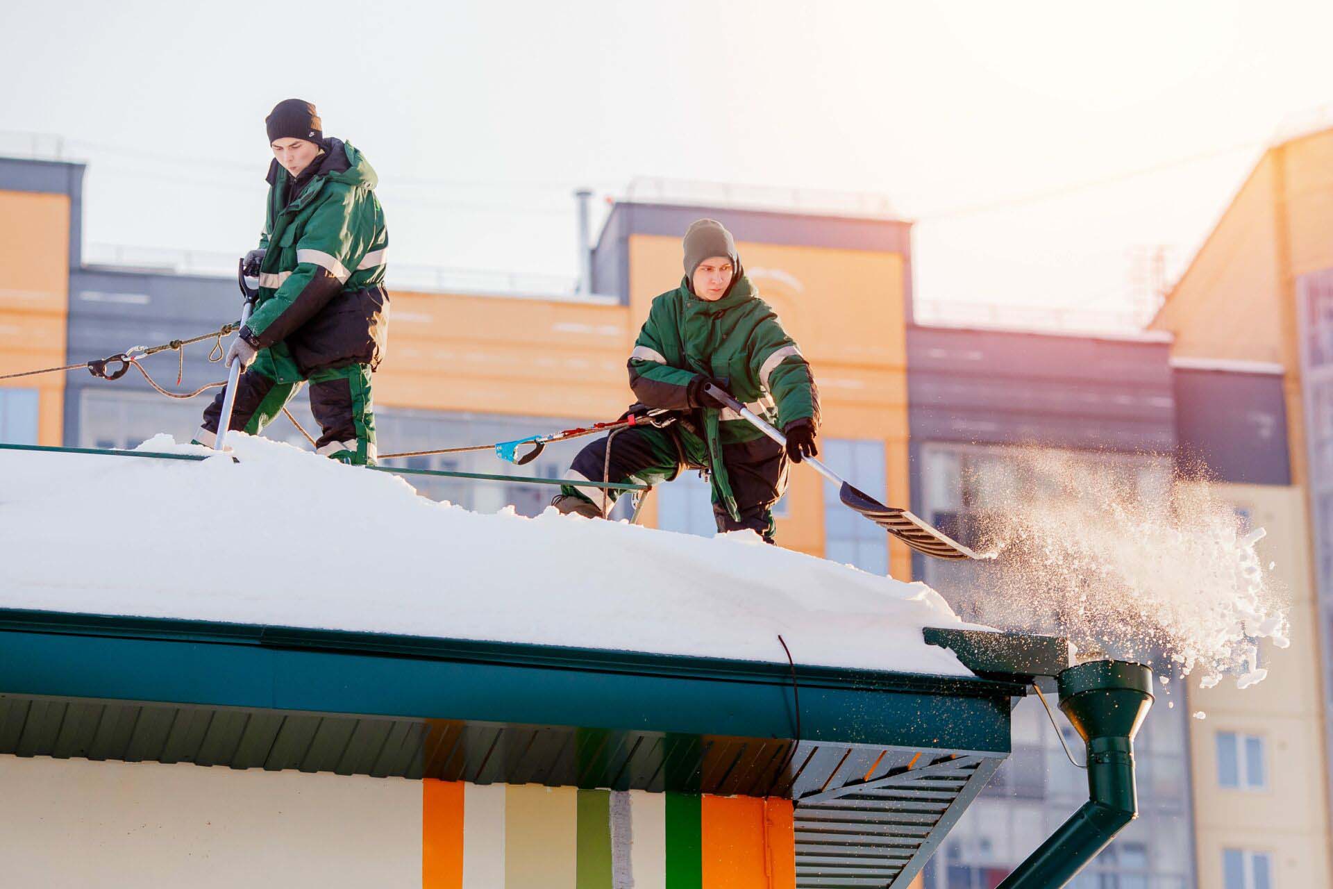 Snow cleaning. Team of male workers clean roof of building from snow with shovels in securing belts of mantra. Insurance for Snow and Ice Removal Businesses