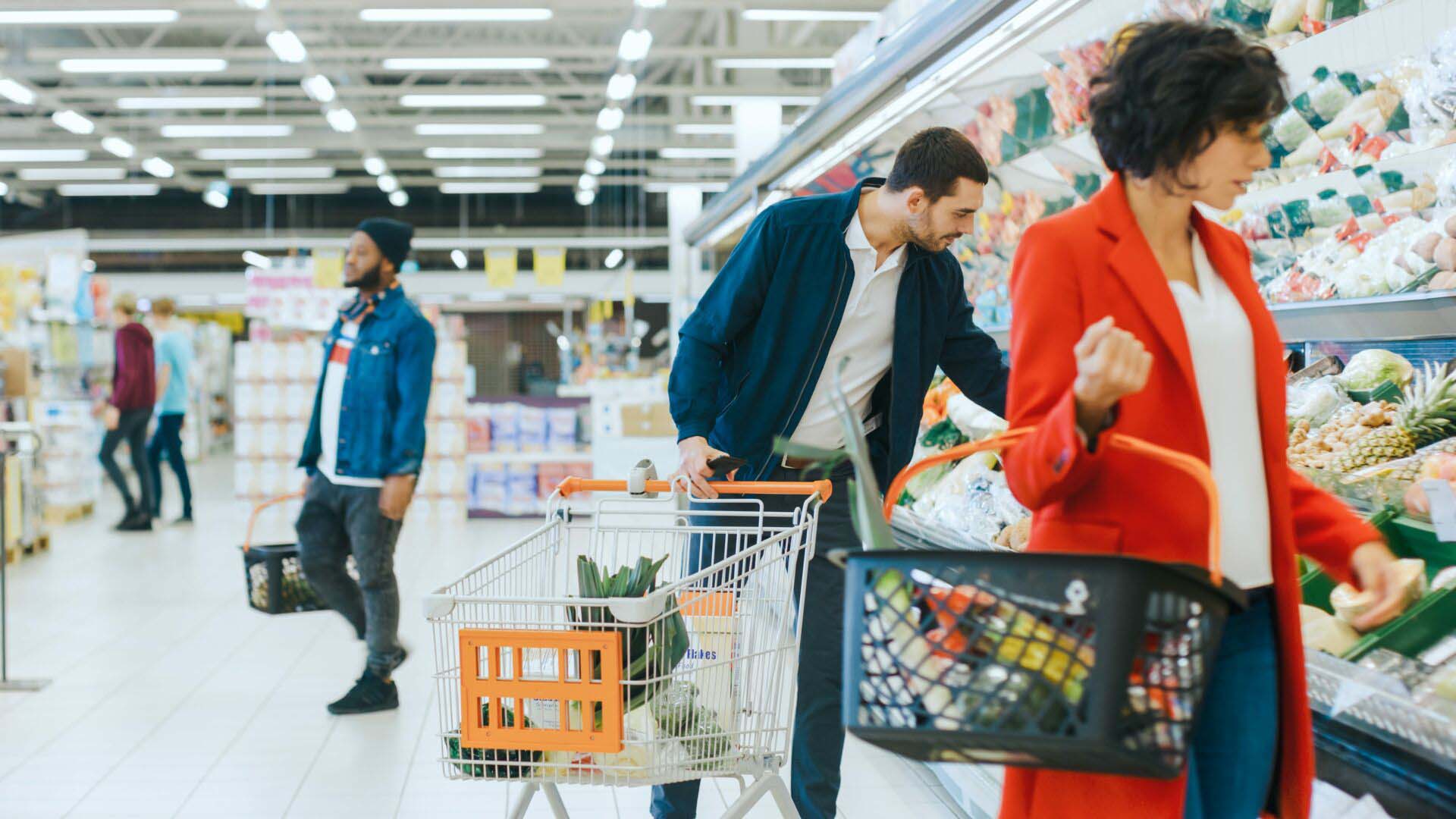 At the Supermarket: Handsome Man with Smartphone, Pushes Shopping Cart, Walks Through Fresh Produce Section of the Store, Chooses Some Products. Other Customers Purchasing Products. Insurance for Grocery Stores