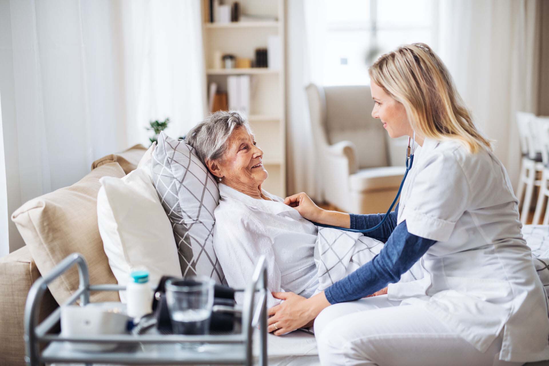 A health visitor examining a sick senior woman lying in bed at home with stethoscope. Insurance for Home Healthcare Providers
