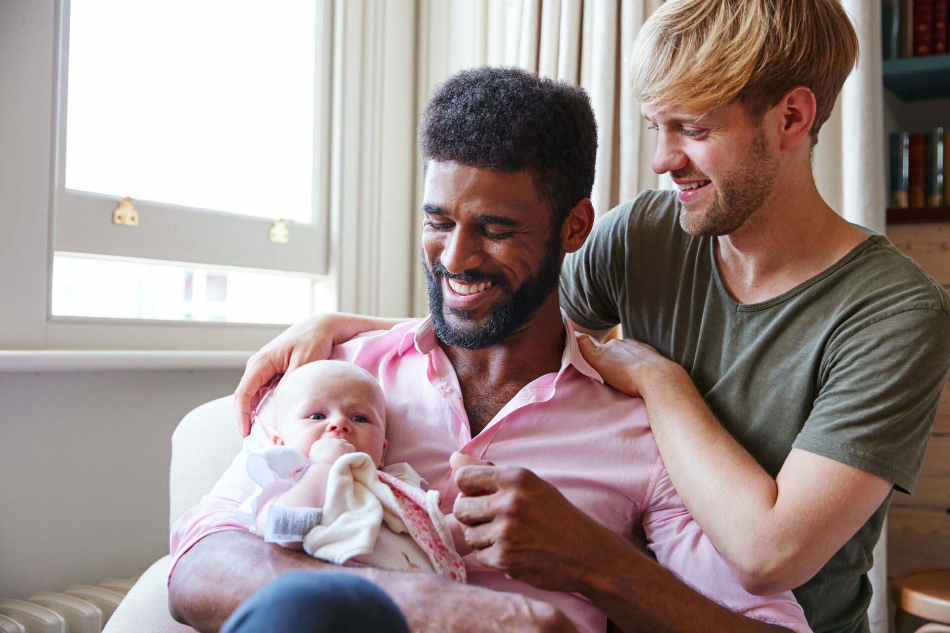Loving Male Same Sex Couple Cuddling Baby Daughter On Sofa At Home Together Insurance for Adoption Agencies