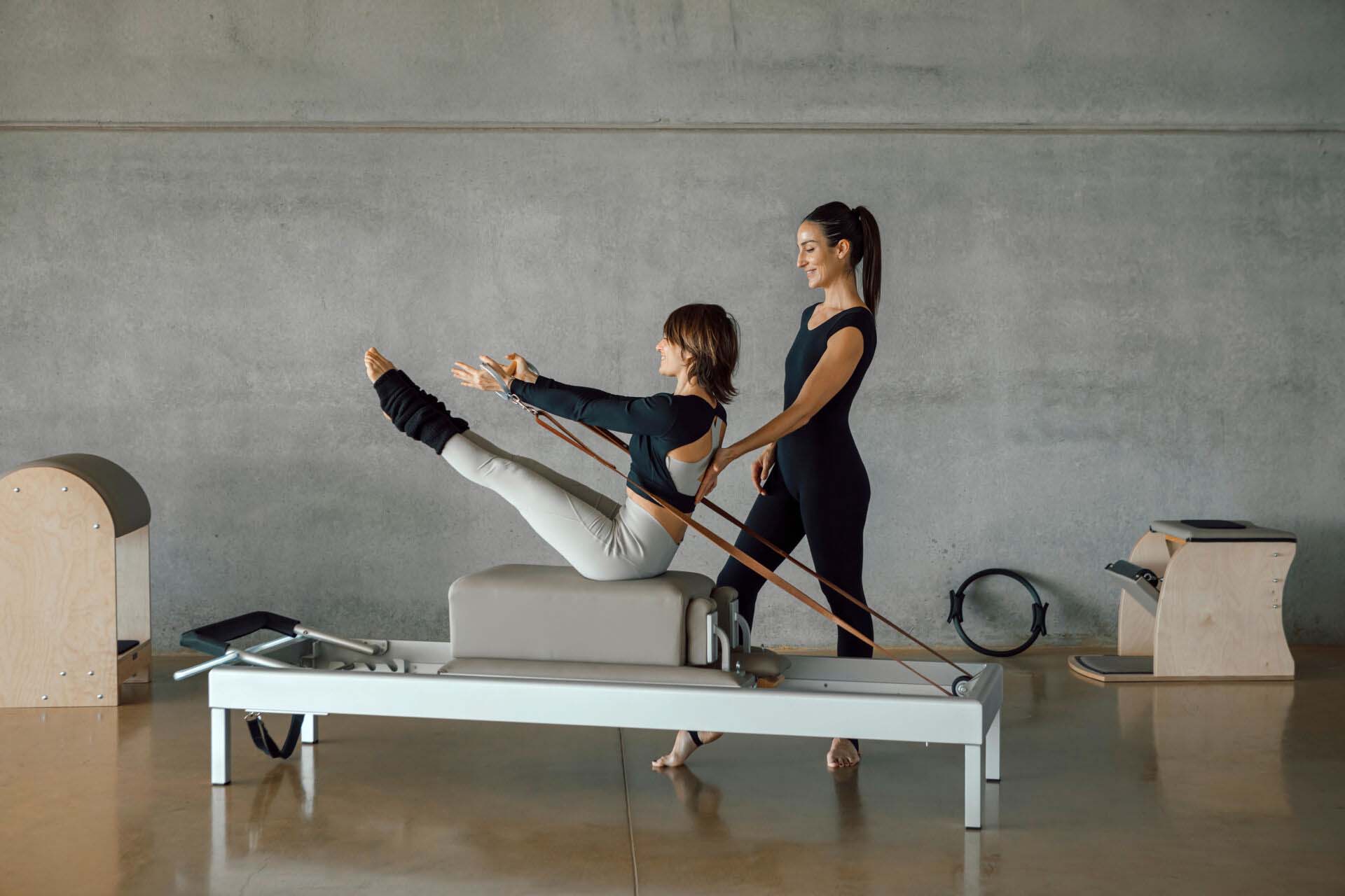 Slim fit Woman doing core exercises while taking Personal Pilates lesson on a Reformer machine, by a female instructor at a big gymnasium center, wide industrial studio. Healthy fitness lifestyle. Insurance for Pilates Instructors