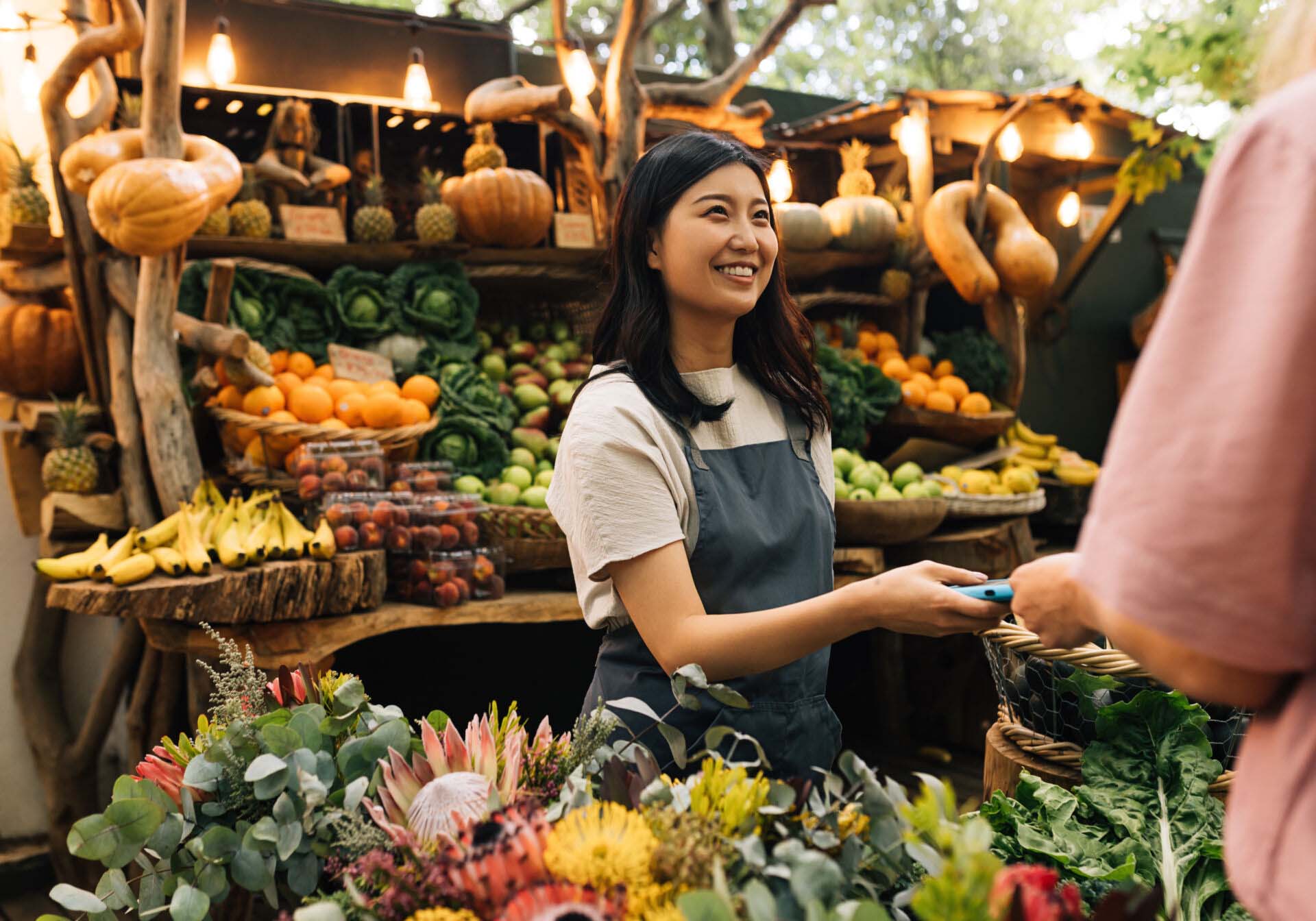 Vendor receiving payment on the outdoor market. Woman in an apron working at a farmers market looking at customer. Insurance for Farmer’s Markets