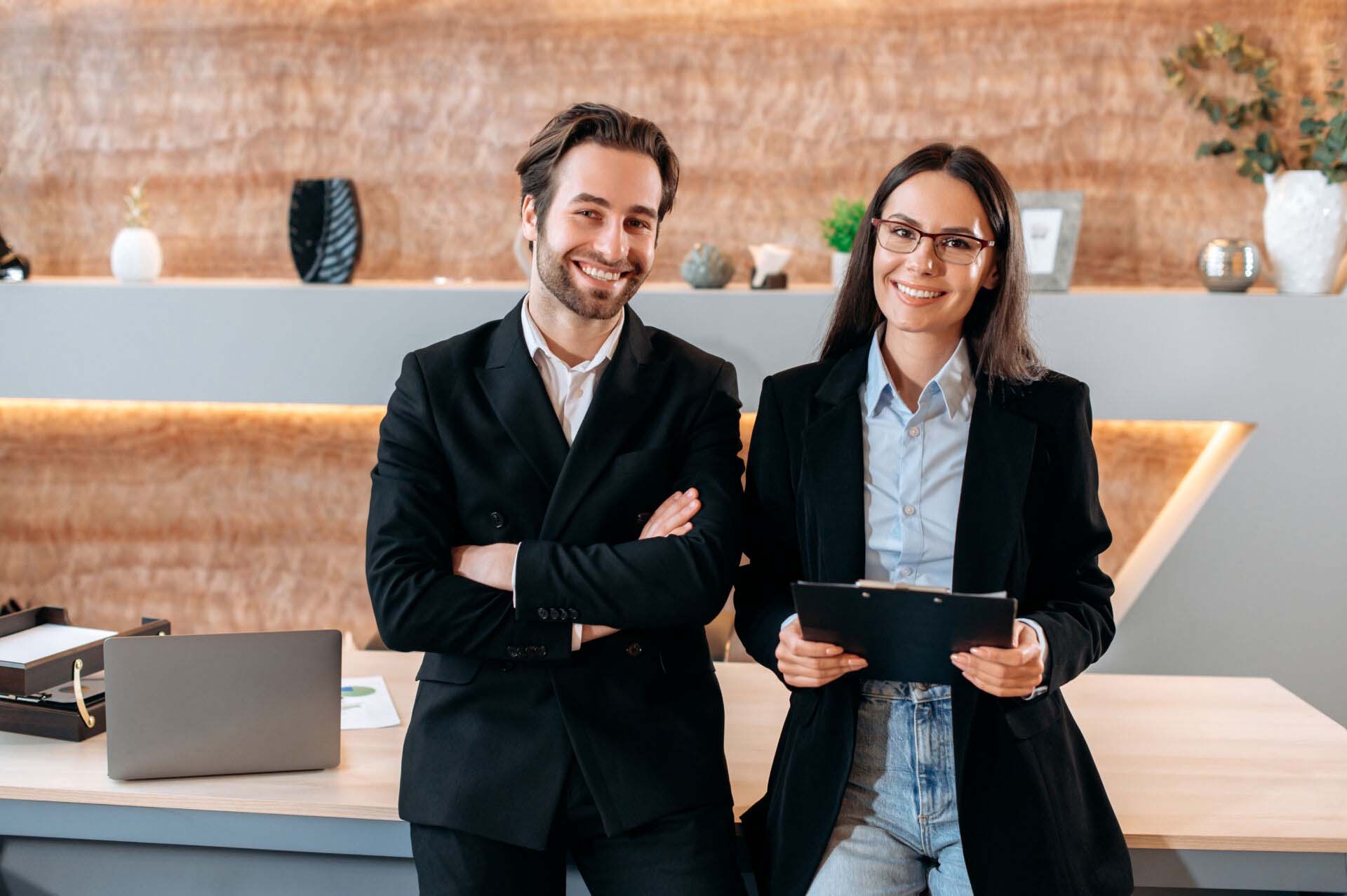 Portrait of two caucasian confident successful company employees, business colleagues, a man and a woman, stand near the desktop in a modern office, look at the camera, smile friendly Insurance for Mortgage Brokers