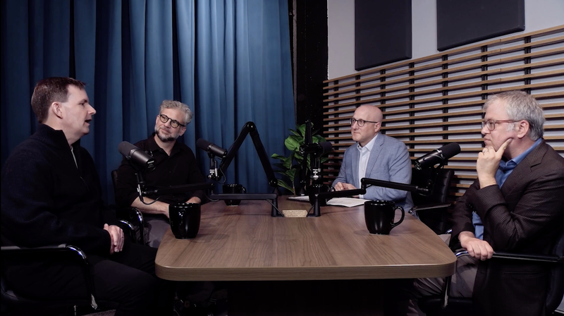 Four men sit around a wooden table with microphones and mugs in a podcast studio setting.