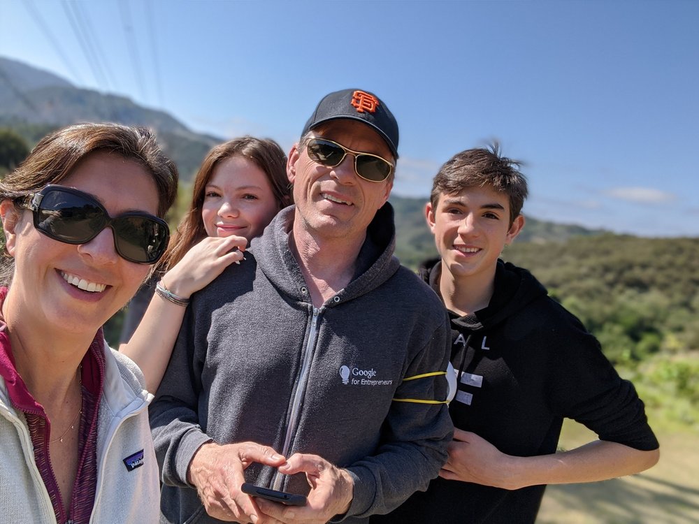 Monica and her three family members are smiling at the camera outside on a sunny day. Monica and her husband are both wearing sunglasses.