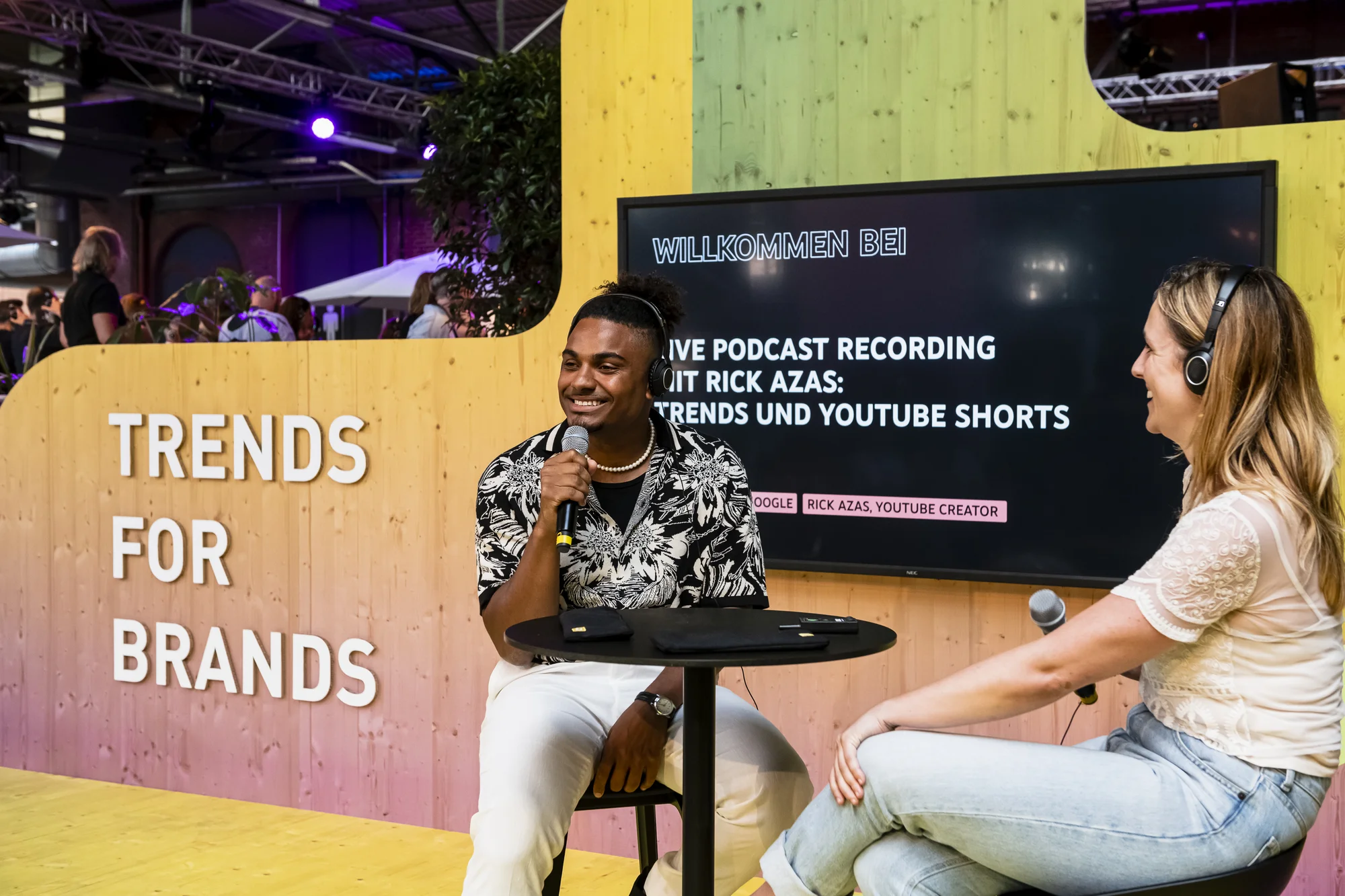 Zwei Personen sitzen auf Hockern an einem Tisch und unterhalten sich. Im Hintergrund ist der Schriftzug "Trends for Brands" zu lesen.