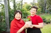 Two women wearing red tops stand in a garden holding a potted plant.