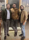 Three men are smiling and standing in a restaurant. The man in the center has his ankles crossed.