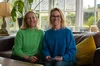 Two women sat indoors on a sofa in a house both in jumpers and smiling.