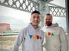 Two men are smiling and standing in front of a window with a city view, wearing Voda-branded hoodies.