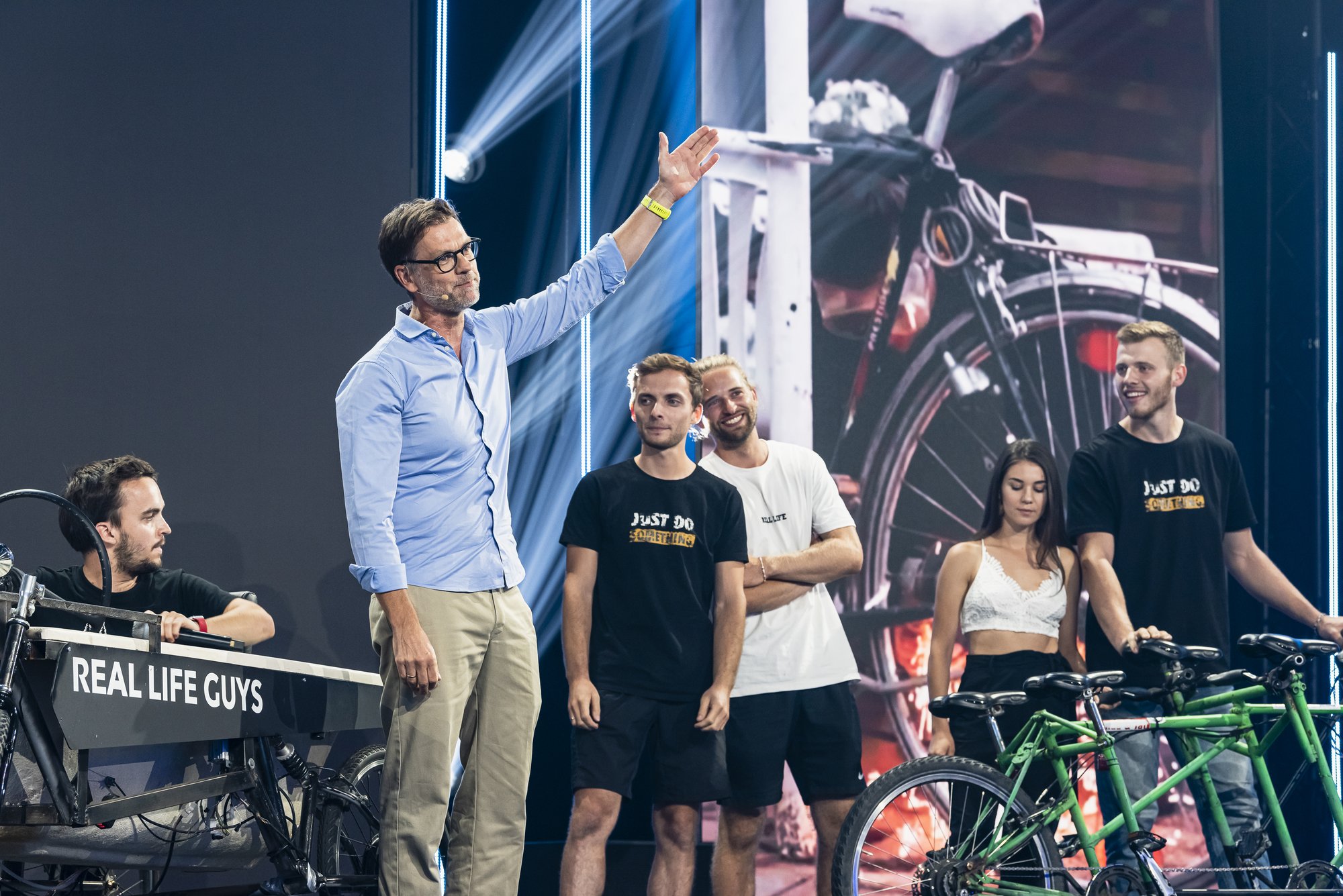 Personen stehen auf einer Bühne, neben ihnen ein Fahrrad auf dem der Schriftzug "Real Life Guys" steht.