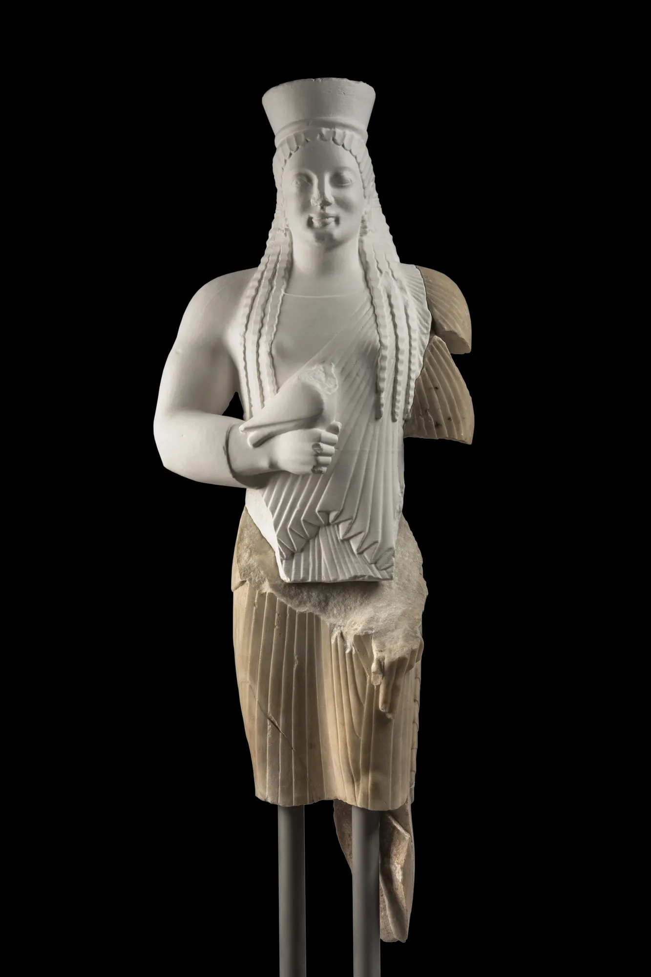 Greek statue of a young woman dating back to around 540 BC. The Kore stands in a traditional pose, wearing a long-sleeved chiton and a short himation draped over her shoulders. She holds an offering in her left hand, which was likely a fruit or flower.