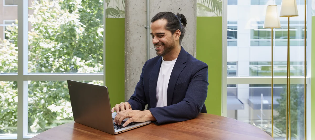 A man in a blazer sits at a wooden desk and uses a laptop computer in an office.