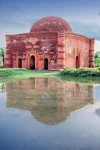 Zdjęcie meczetu Chunakhola w Bagerhat w Bangladeszu. Meczet stoi na zielonych polach uprawnych. Na pierwszym planie widać odbicie meczetu w kałuży wody.