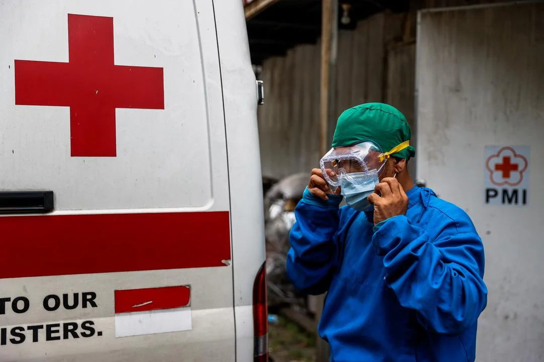 A photo of medical personnel in protective clothing and goggles next to the back of Red Cross van