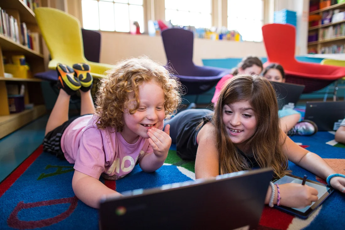 two smiling children in a classroom looking at an educational laptop