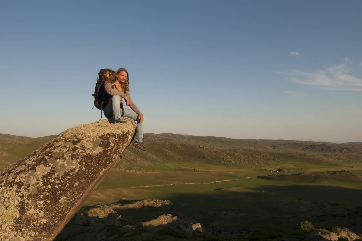 A photograph of Gino Caspari sitting on a rock outdoors. There is a sloping mountain range in the background.