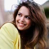 A woman with long brown hair and wearing a yellow blazer looks over her shoulder, smiling.