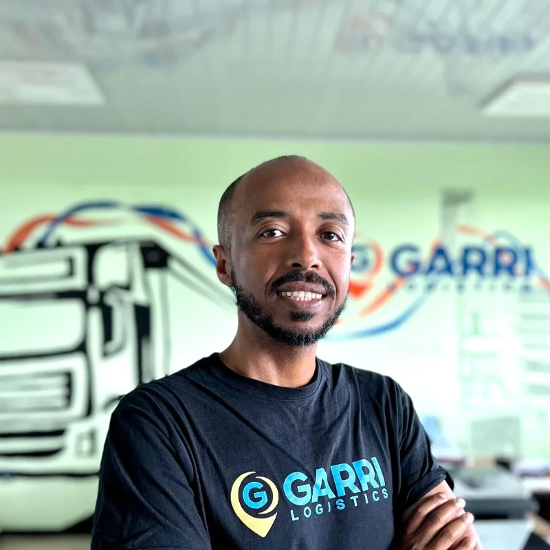 A Black man with facial hair stands with his arms folded, looking at the camera. He's wearing a black t-shirt that says "Garri Logistics" in blue across the front, and there's a mural of a truck on the wall behind him.