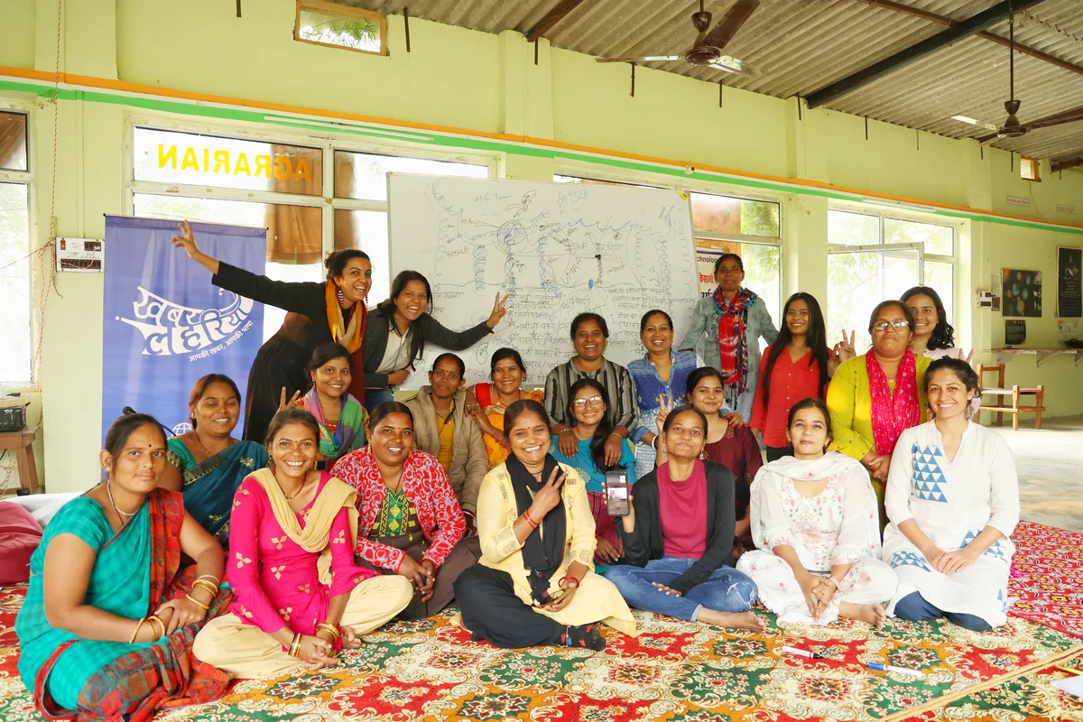 A group of 21 women mostly wearing brightly coloured saris. 18 of them are seated in 2 rows with the remaining 3 standing behind them giving a freedom salute with their first two fingers.