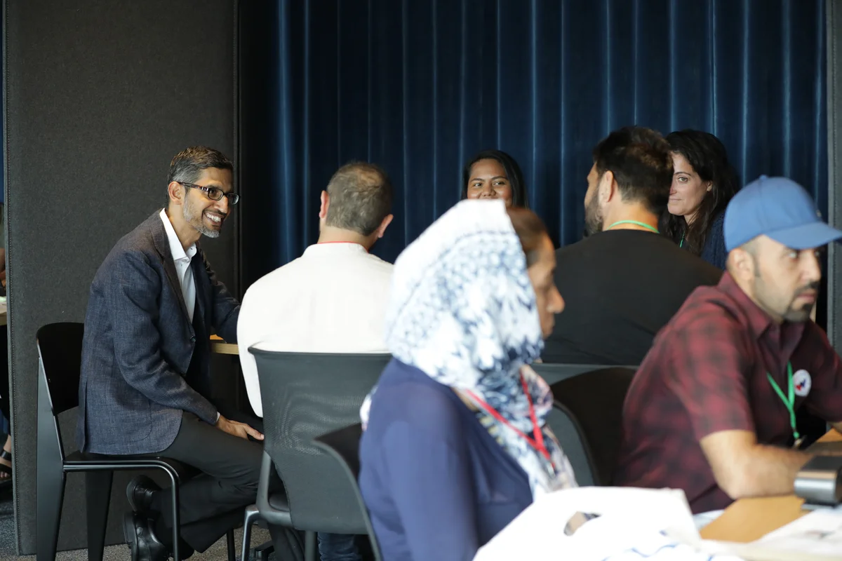 Sundar Pichai seated a table with four other people. He's smiling and speaking with them. In the foreground are two people at another table.