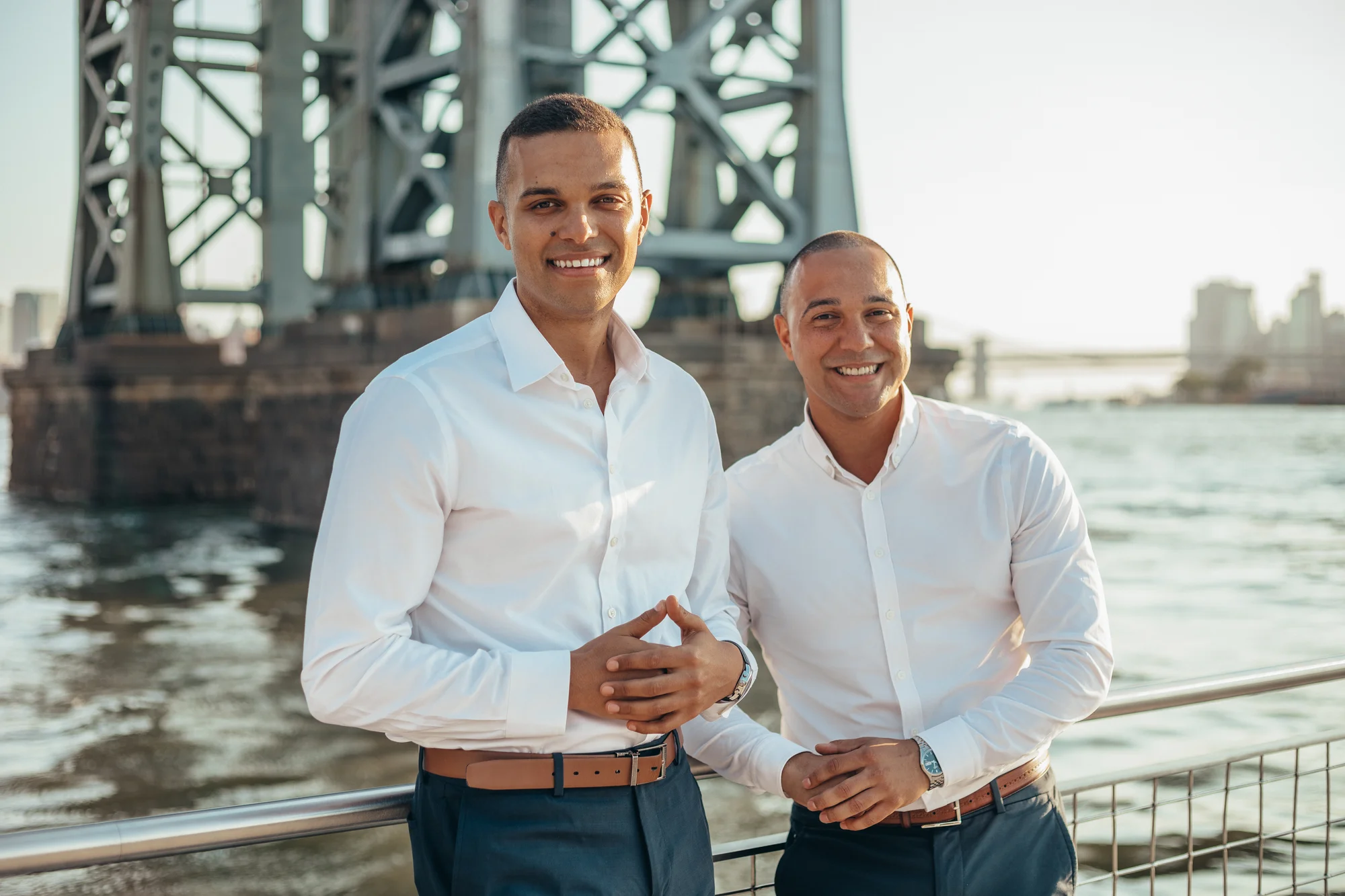 Team photo of Brian Hollins and Aaron Samuels, both wearing long-sleeved white button down shirts, brown belts, and navy slacks.  They are standing in front of a river.