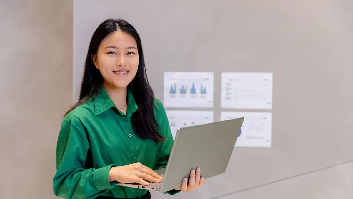 A young female in green shirt is smiling, while holding a laptop. Behind her are images of charts and graphs on the wall.