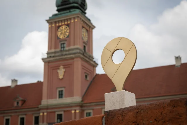Na pierwszym planie nagroda „Złota Szpilka”. Za nią wieża zegarowa z czerwonej cegły Zamku Królewskiego w Warszawie.