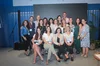 A group of 20 people , mostly women and all wearing lanyards, smile at the camera in front of a gray step-and-repeat. Those in the back are standing while four women in the front sit on clear plastic chairs.