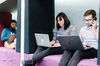 Three people working on laptops while sitting on a purple cushion, divided by a white wall. One person is using a cell phone.