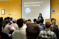 Carrie stands in front of a group of people speaking while holding a microphone. A presentation behind her reads: “Neural substrates of Musical Creativity.”