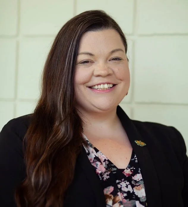 Image showing a person looking into the camera and smiling. The person is wearing a black blazer.