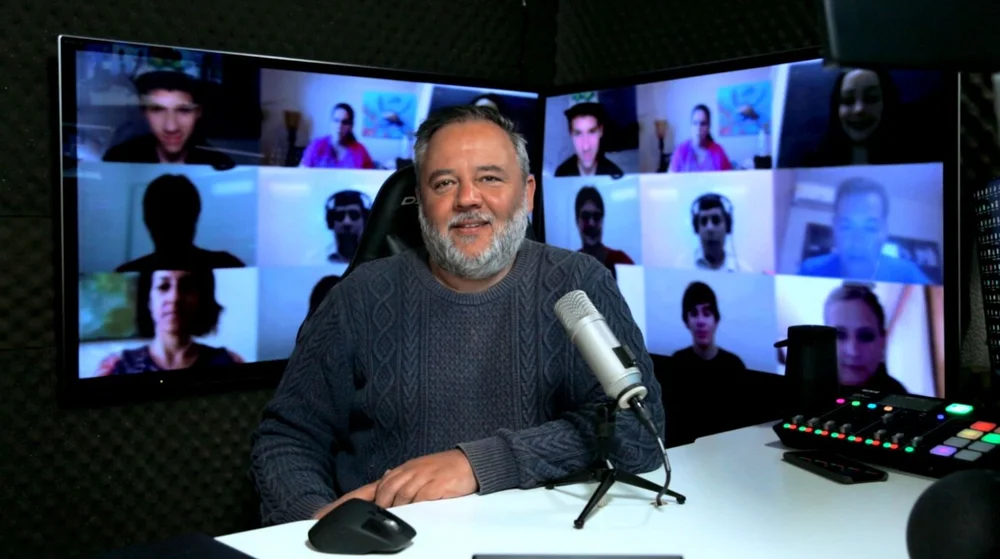 Miguel, wearing a dark blue knitted sweater, is sitting at a desk in front of a gray microphone and smiling at the camera. In the background are two screens showing many people on a video call.