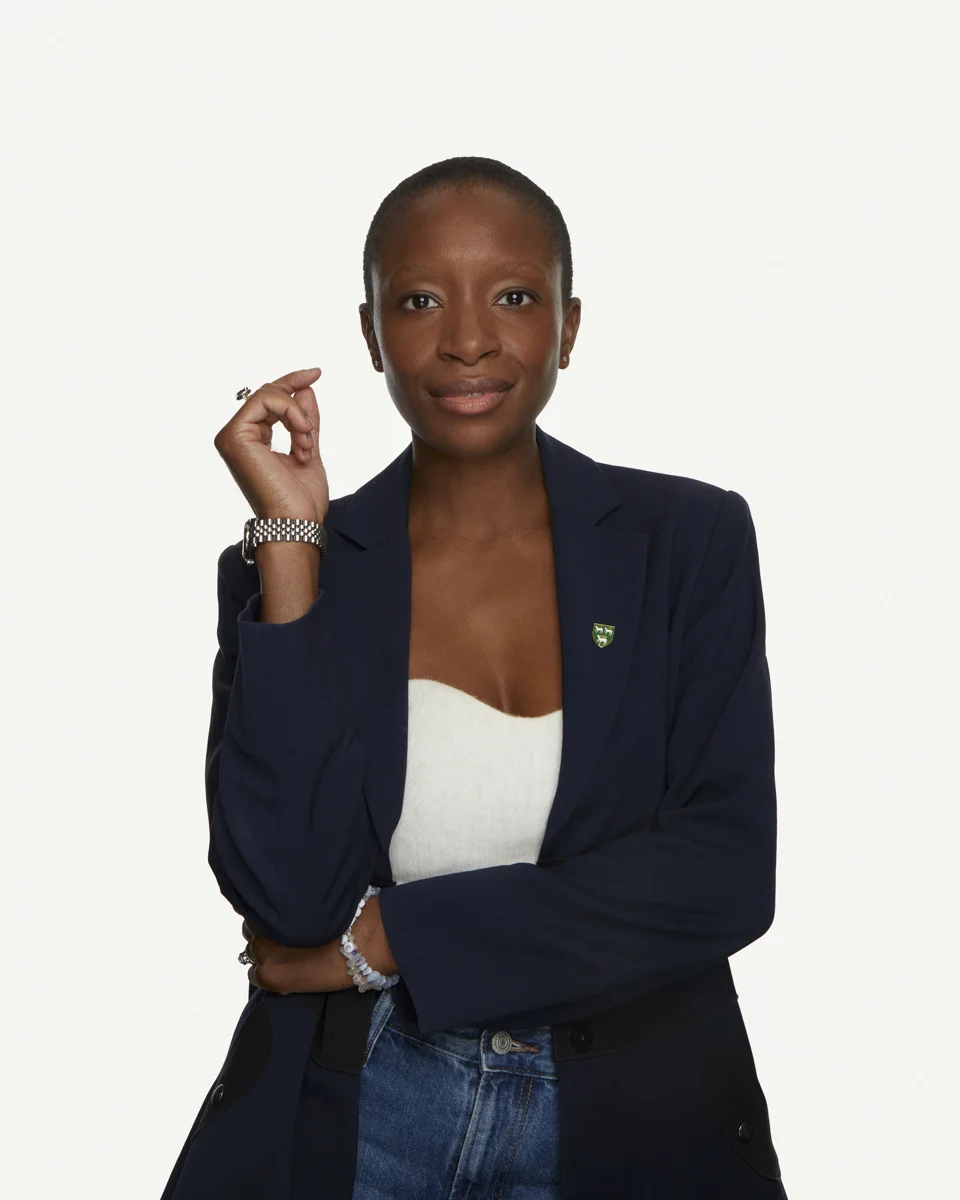 A Black woman faces the camera in front of a white background, wearing a black blazer, white top and blue jeans. One arm is folded across her torso, propping up her other arm vertically.