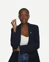 A Black woman faces the camera in front of a white background, wearing a black blazer, white top and blue jeans. One arm is folded across her torso, propping up her other arm vertically.