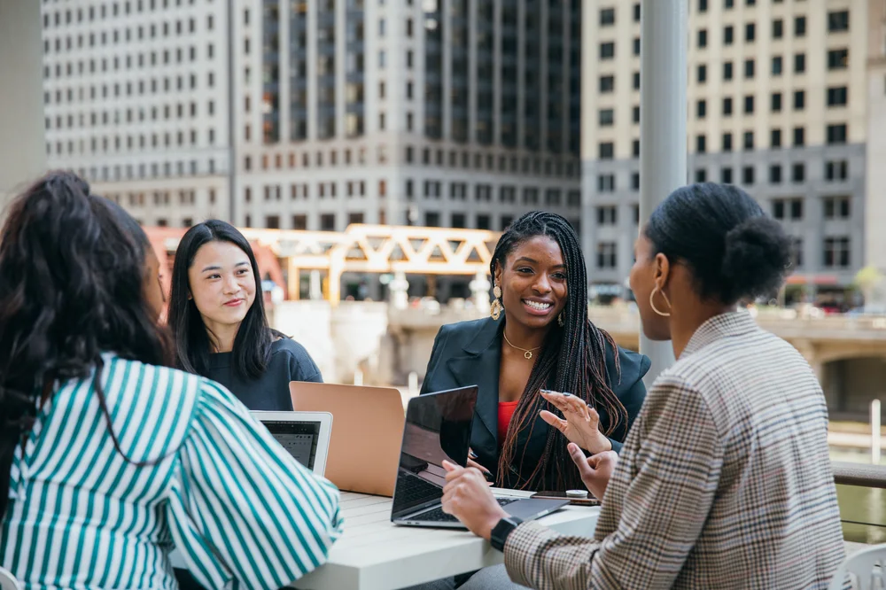 Four women in professional attire sit at a table outdoors conducting a meeting
